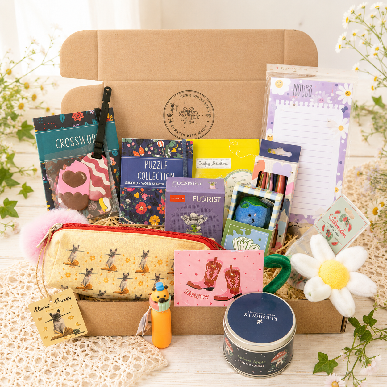 A curated gift box filled with stationery, puzzles, a pencil case, a candle, and small accessories, arranged on a white lace surface with flowers around it. Visible items include a brown kraft gift box, a patterned yellow pencil case, a pink notepad, a puzzle book, a crossword card, and a round tin candle. A curated mystery scoop from Down Whimsy Way.