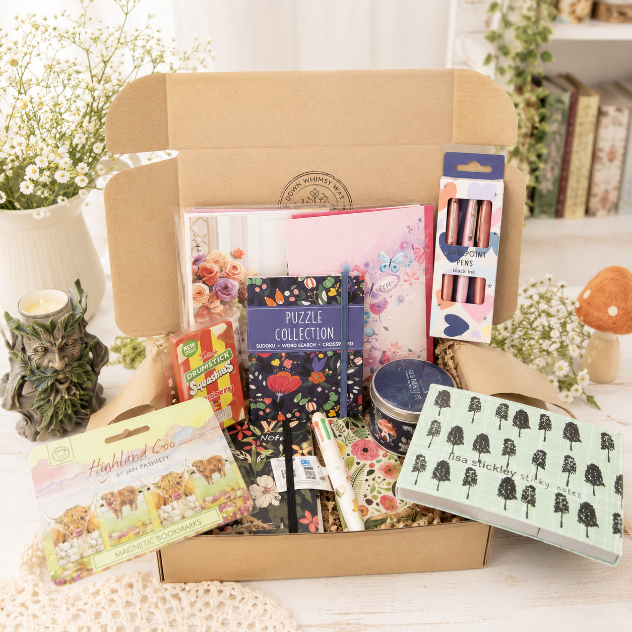 A Curious Explorer curated gift box filled with assorted stationery, craft, and hobby items arranged on a light tabletop. Visible contents include patterned notebooks or cards, a dark blue puzzle book, a pink floral item, a boxed set of metallic pens or pencils, a small card reading 'Highland Cows,' a round tin, and a mint green box with black tree illustrations.
