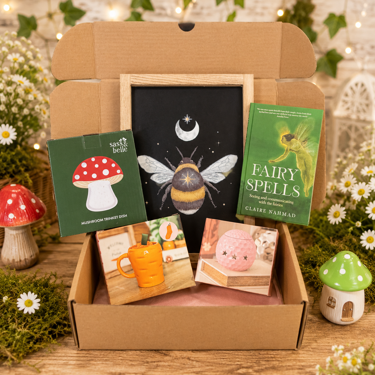 A curated gift box set displayed in an open cardboard box on a wooden surface, surrounded by fairy garden props like small mushrooms, white flowers, and a green 'Fairy Spells' book by Claire Nabrad. The set includes a framed blackboard with a bee and moon illustration, a red-and-white mushroom card, a mug photo, a pink candle photo, and another green book.