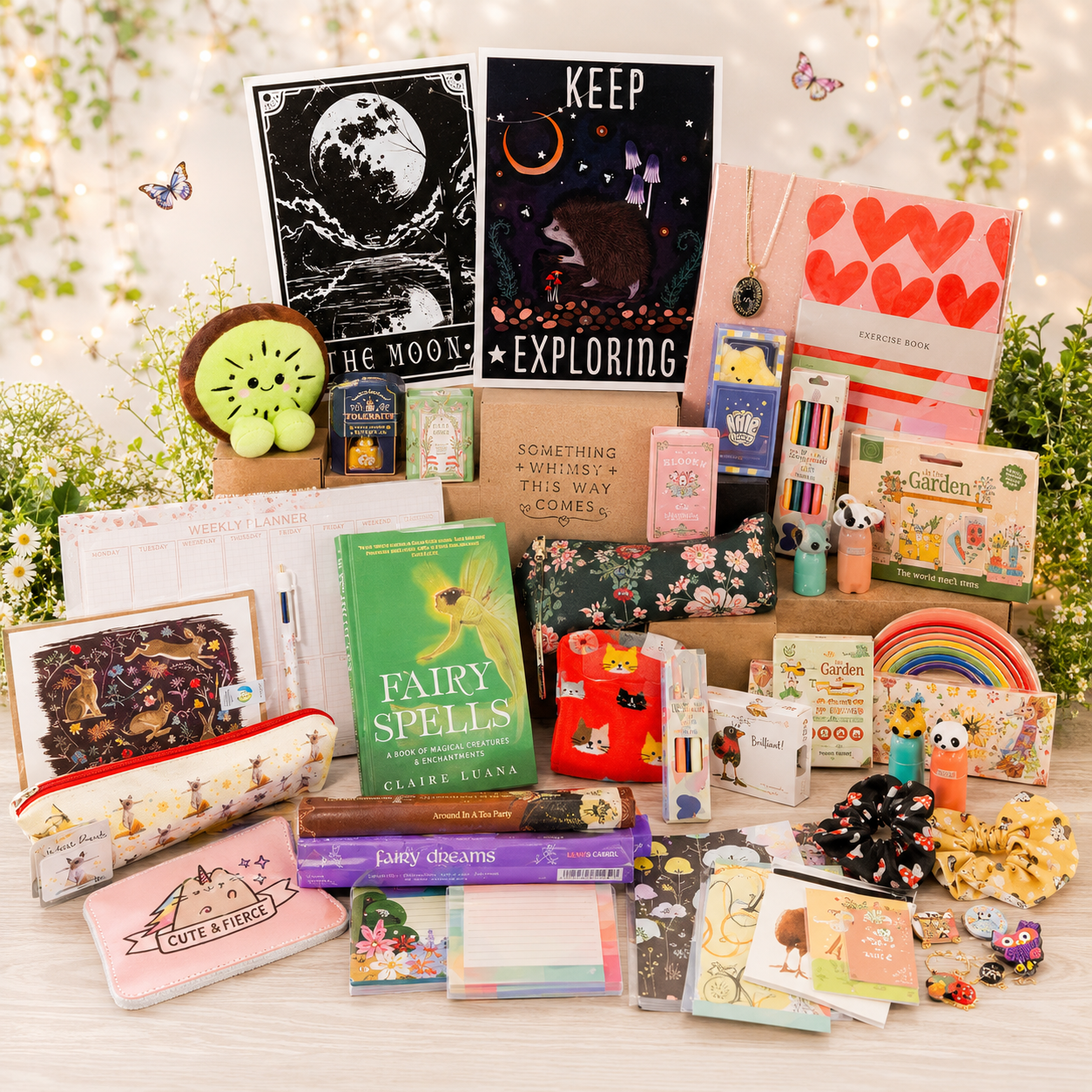A styled flat-lay display of a curated gift basket or bundle containing various cute, whimsical items including notebooks, stickers, a pencil case, a green 'Fairy Spells' book, a small plush, and stationery, arranged on a white surface with decorative flowers and fairy lights in the background.